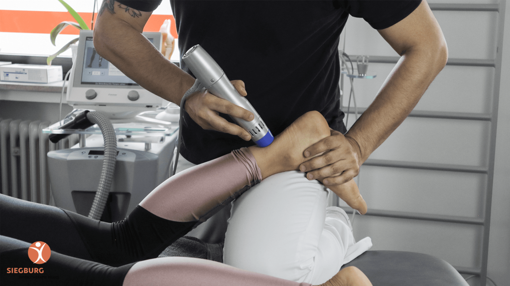 Stoßwellentherapie am Fuß einer liegenden Patientin durch einen Therapeuten in der Praxis